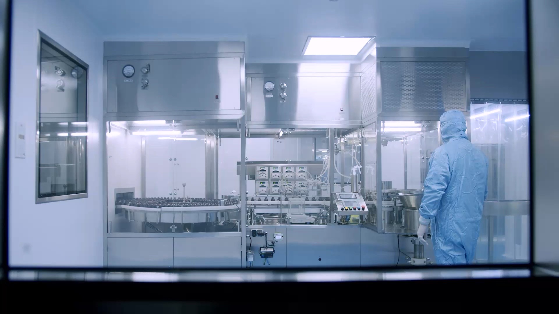 An operator in a cleanroom suit beside an aseptic filling line, seen through a viewing window