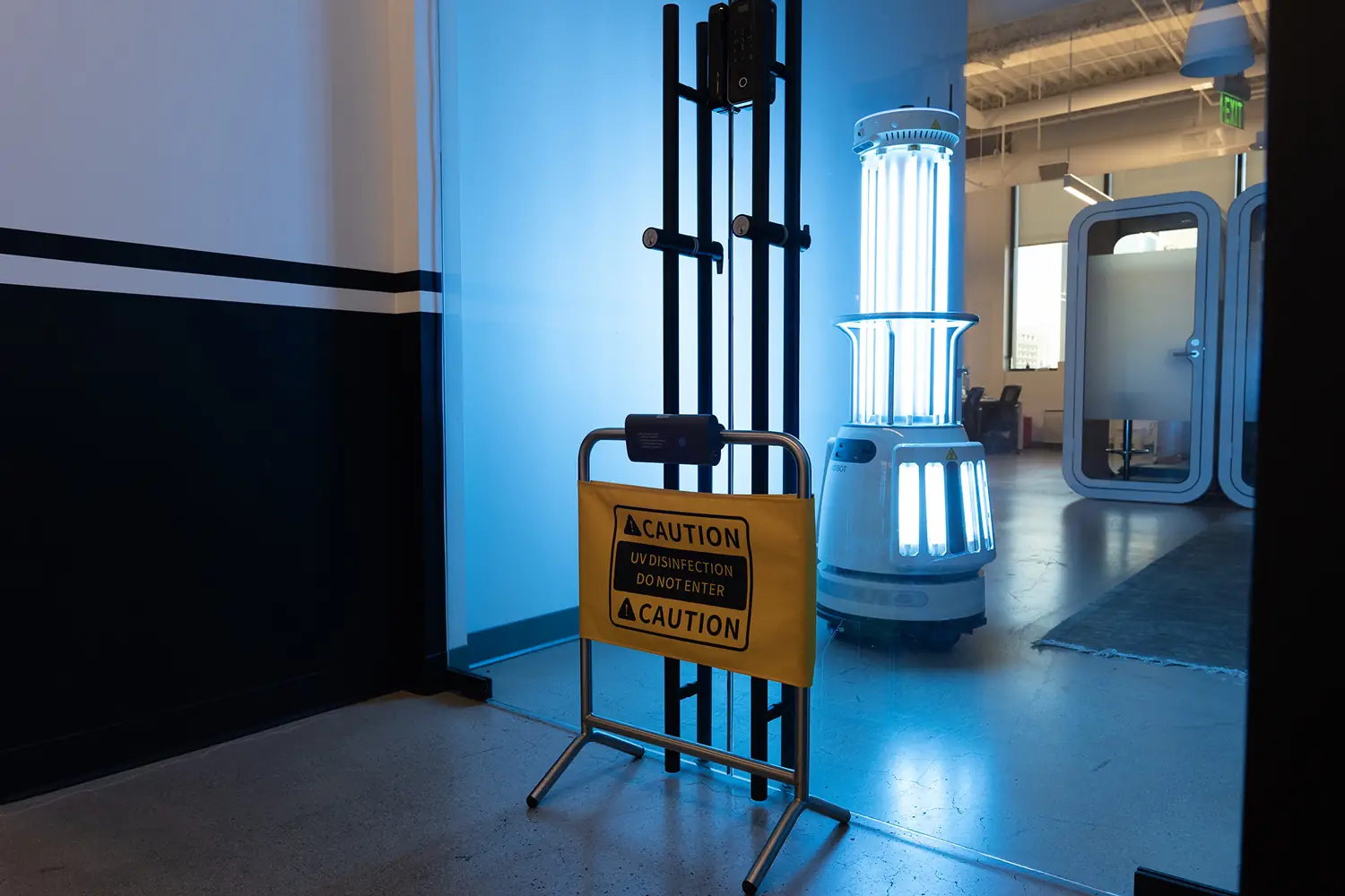 Yellow caution barrier reading “UV disinfection — do not enter” placed across a doorway, with the robot’s lamps lit in the room beyond.