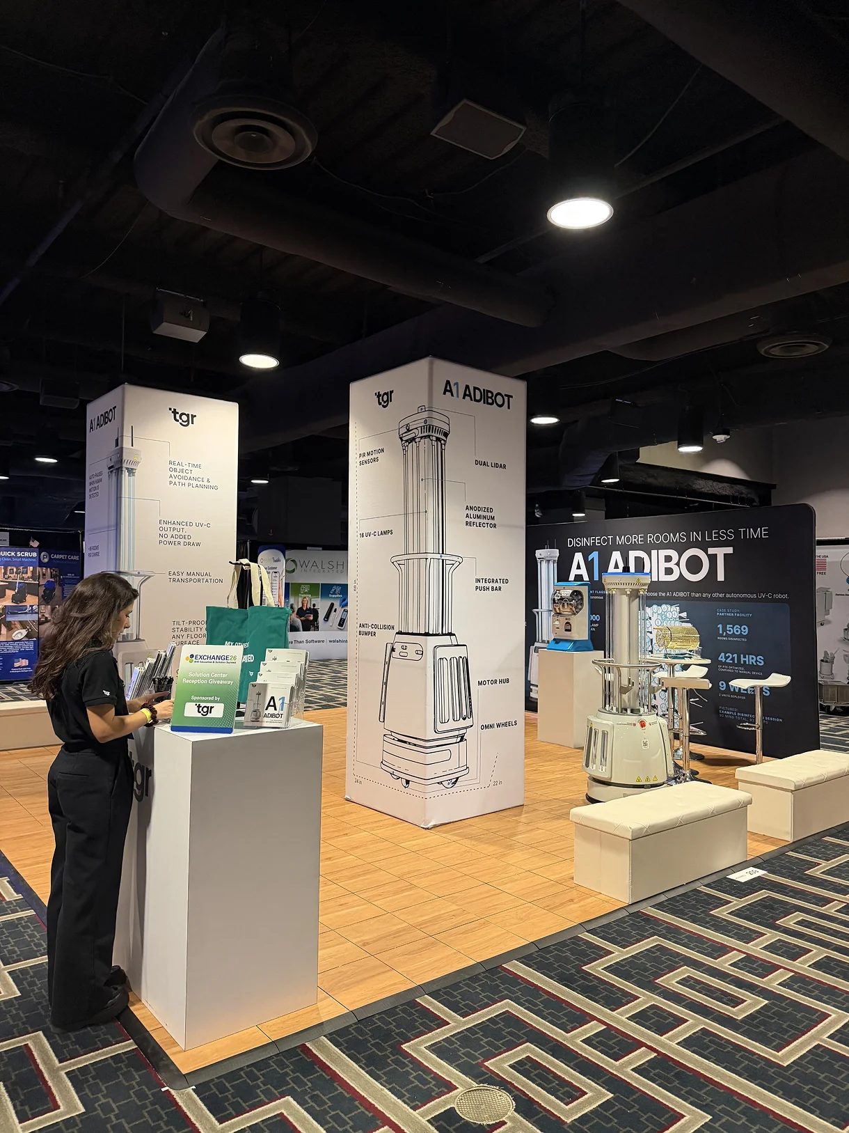 TGR exhibition booth with a tall pillar graphic showing a labeled line drawing of the A1 ADIBOT, a staff member arranging literature on a counter.