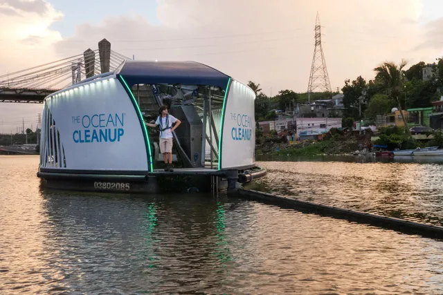 The Ocean Cleanup Interceptor floating beside a riverbank, with a barrier guiding debris toward the vessel.