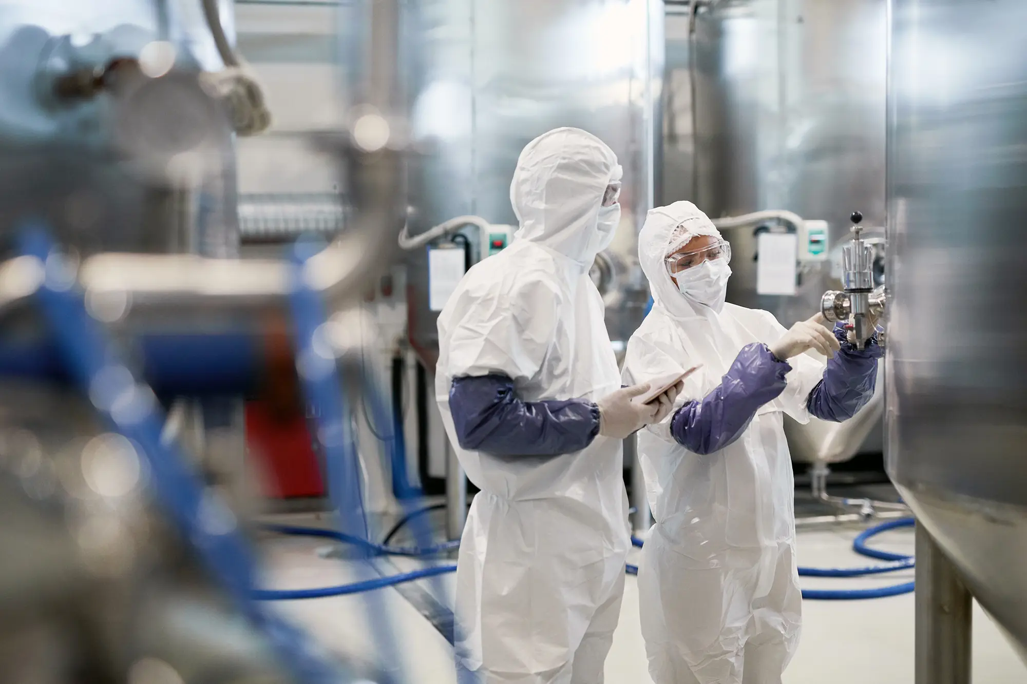 Two workers in full protective garments inspecting equipment in a processing area.
