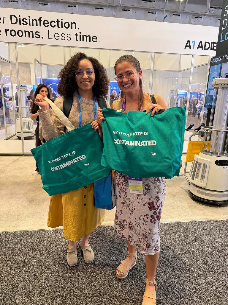 Two attendees holding green TGR tote bags in front of the exhibition booth.