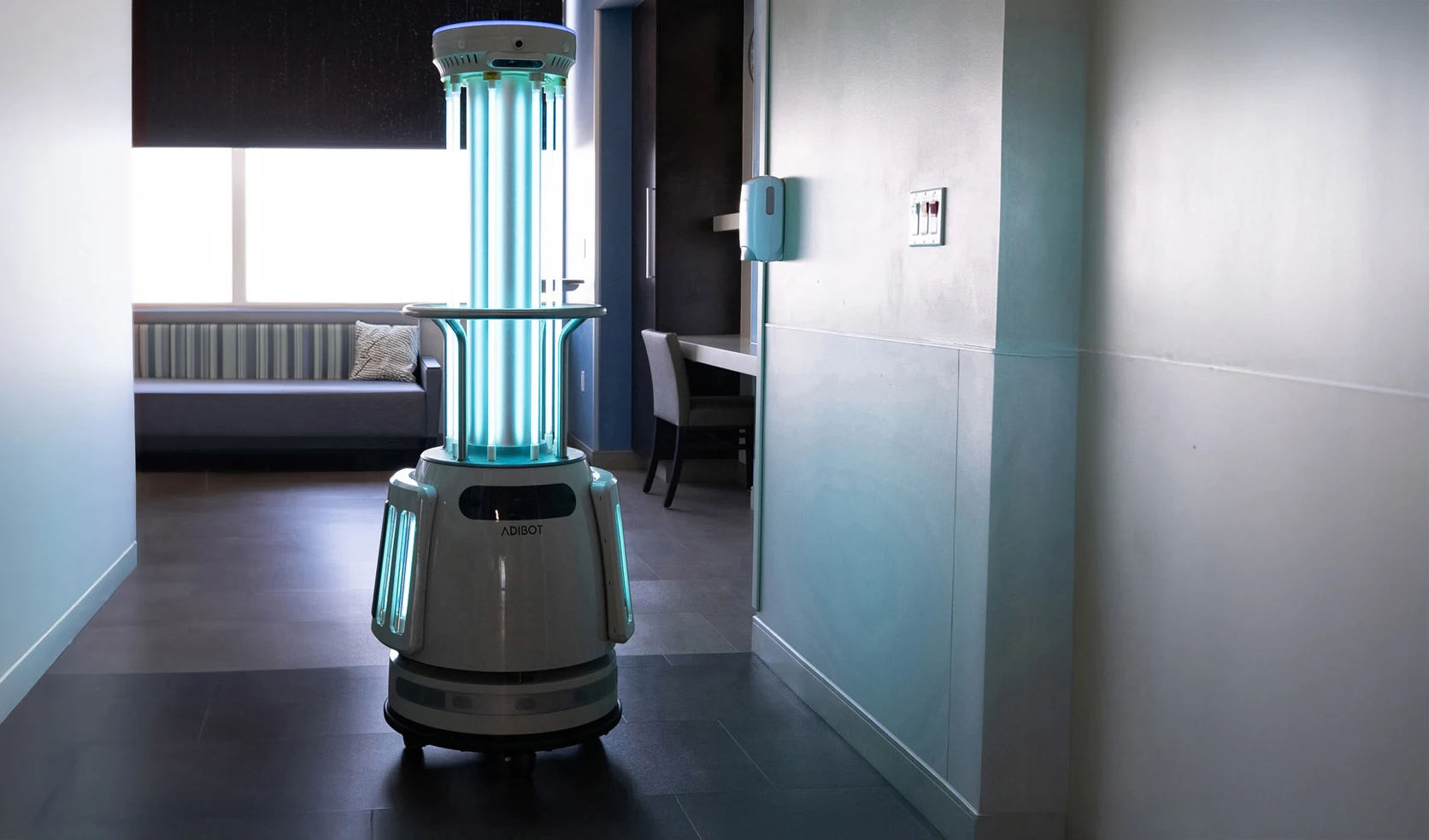 A1 ADIBOT with illuminated UV-C lamps in an unoccupied room beside a wall-mounted dispenser