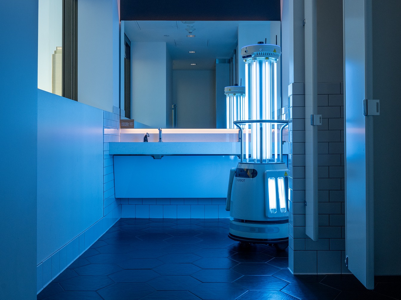 An A1 ADIBOT running its UV-C lamps in a tiled restroom, casting blue light across the wash basins and floor